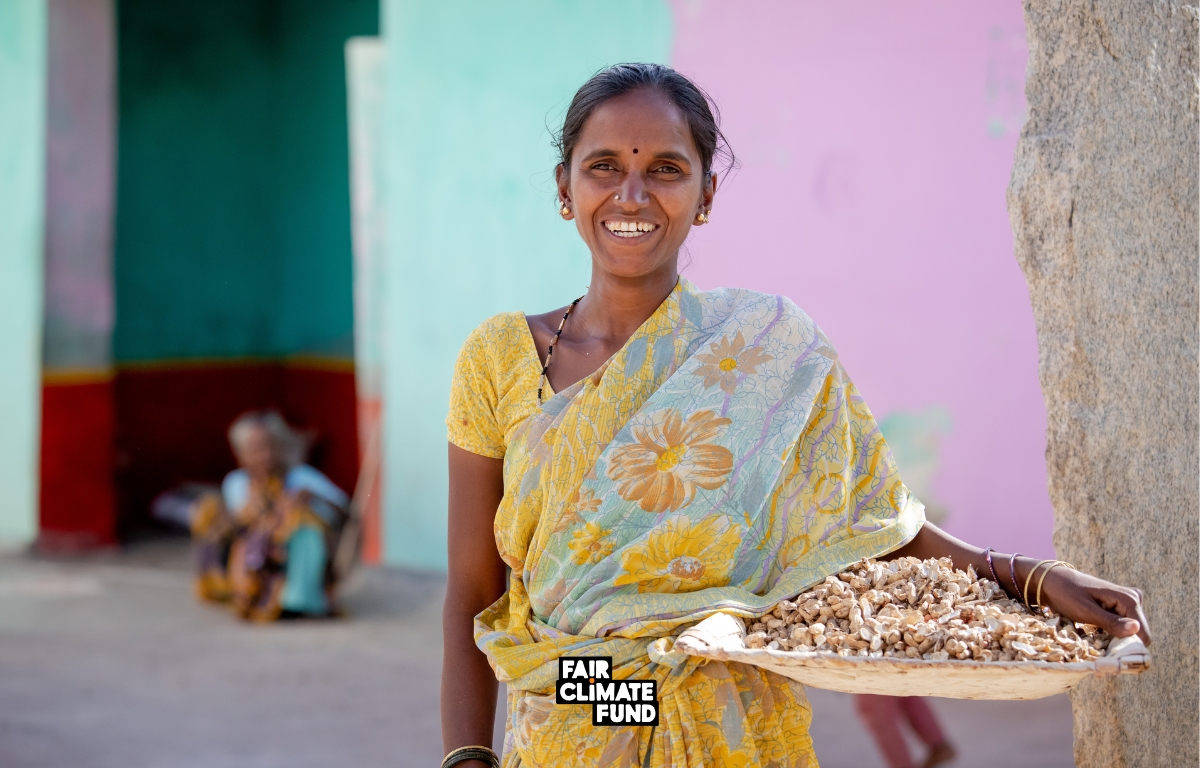 A woman in a yellow sari smiles, holding a woven tray with nuts. A person sits in the background against a pastel-colored wall. "Fair Climate Fund" is printed on the image. Designed by Circular Design, a not-for-profit web design agency.