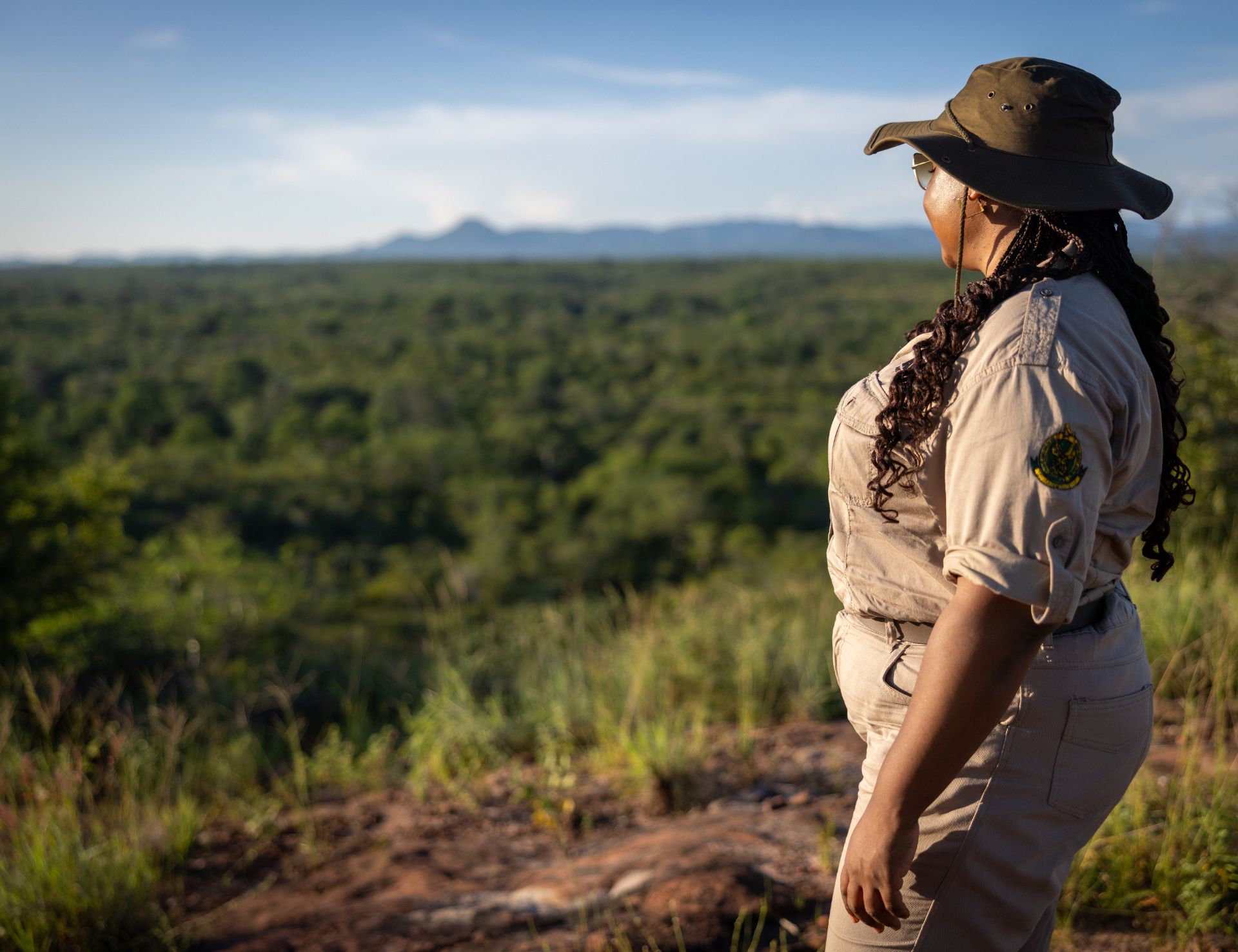 A person in a safari uniform and hat stands on a rocky hill, looking out over a vast, green landscape under a clear blue sky. Designed by Circular Design, a not-for-profit web design agency.