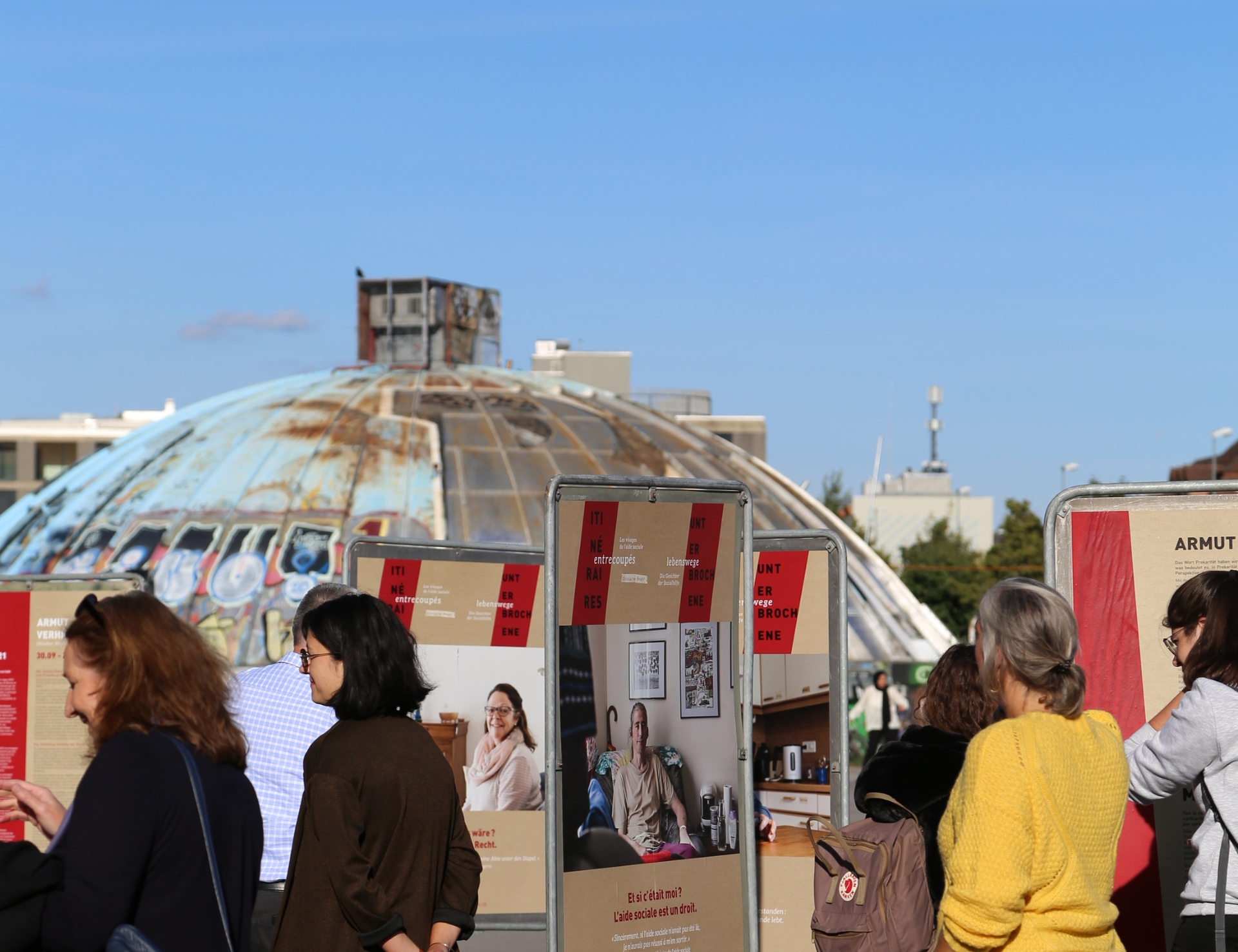 People standing and looking at outdoor display boards with a weathered, dome-shaped structure in the background under a clear blue sky. Designed by Circular Design, a not-for-profit web design agency.
