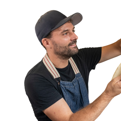 Man in a black t-shirt, denim apron, and cap stretching dough with both hands, facing to the right. Designed by Circular Design, a not-for-profit web design agency.