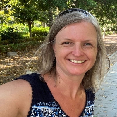 A woman with shoulder-length blonde hair is smiling outdoors, standing on a paved path with trees and greenery in the background. Designed by Circular Design, a not-for-profit web design agency.