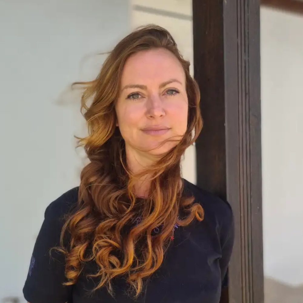 A woman with long, wavy auburn hair stands in front of a white wall and a dark wooden beam, looking at the camera with a neutral expression. Designed by Circular Design, a not-for-profit web design agency.