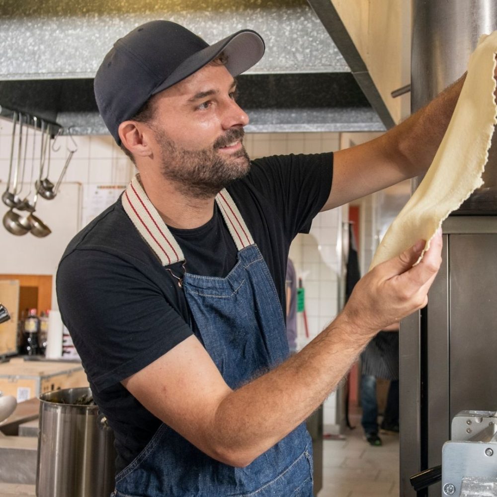 A man in a blue apron and cap stretches dough in a commercial kitchen, with metal utensils hanging in the background. Designed by Circular Design, a not-for-profit web design agency.