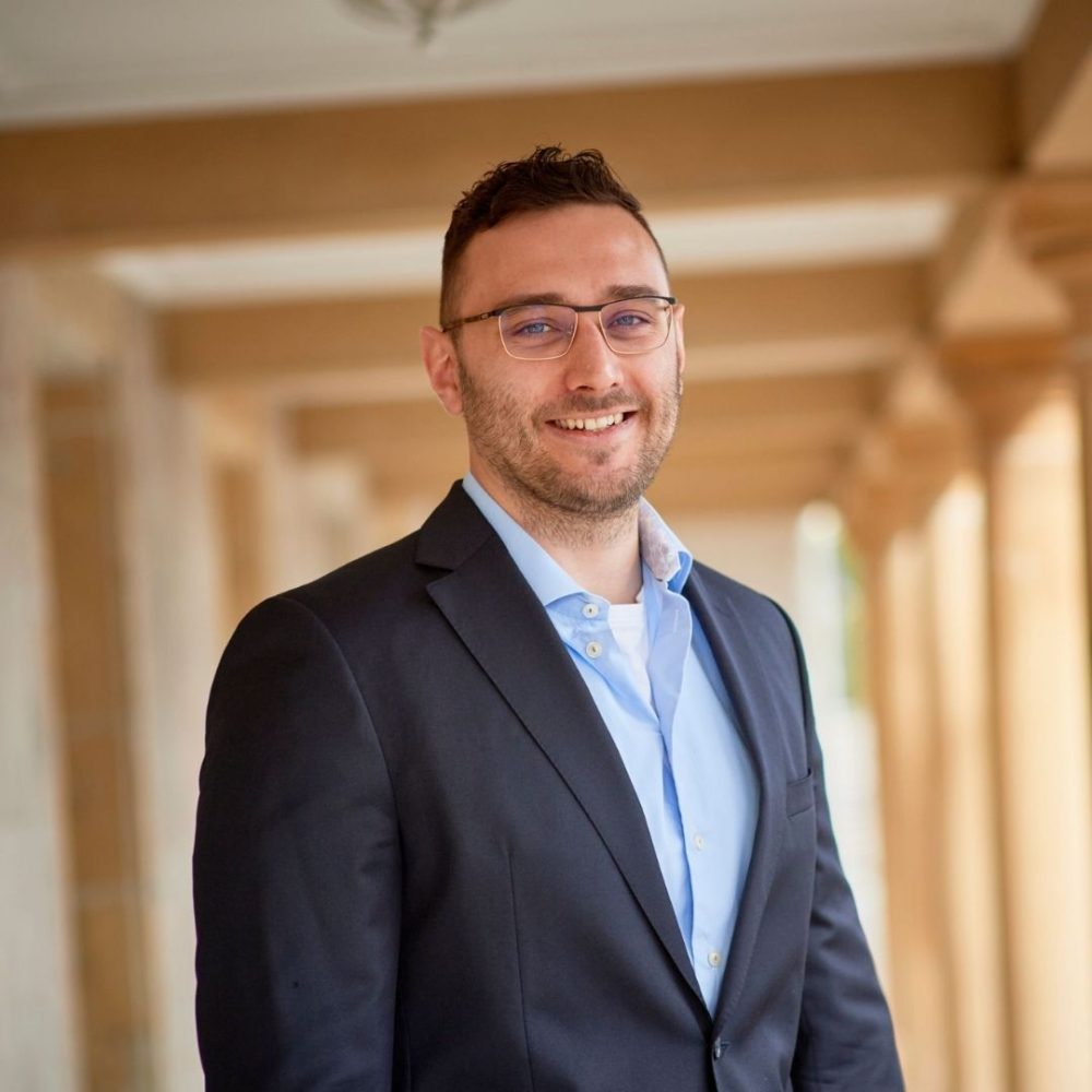A man wearing glasses and a dark suit jacket over a light blue shirt stands smiling in an outdoor corridor with columns in the background. Designed by Circular Design, a not-for-profit web design agency.