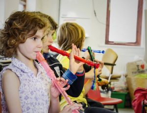 Children sitting in a row play recorders in a classroom setting.