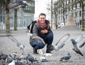 Susanne Clauss crouching among pigeons in an urban area, smiling, wearing glasses and a backpack, surrounded by trees and buildings.