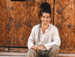 Person with long dreadlocks sitting and smiling in front of a wooden door, wearing a light shirt and pants.