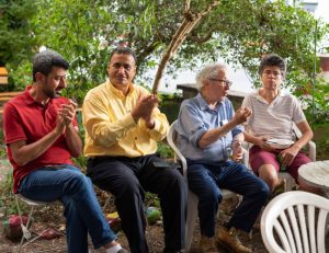 Four people sit together outdoors on chairs, with trees in the background. Two are clapping, one is holding a cup, and one is holding a plate with food.