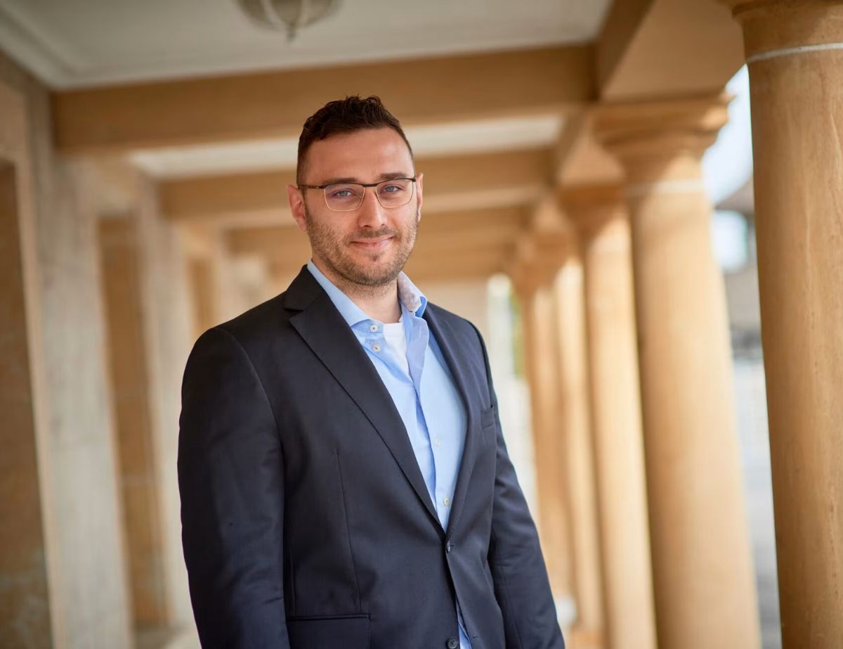 A man in a suit stands in a corridor with columns, looking at the camera and smiling slightly. Designed by Circular Design, a not-for-profit web design agency.
